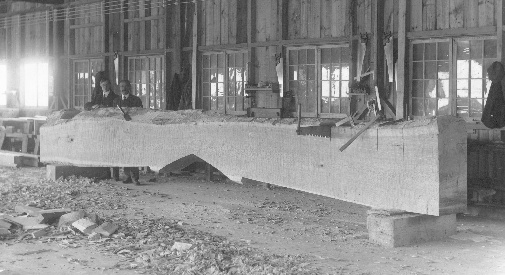 Raymond Pitcairn and Harry Bowman stand by a tie beam taking form out of a rough-hewn log. Bowman, a master carpenter, was in charge of the wood working operation, and later headed the entire building force.