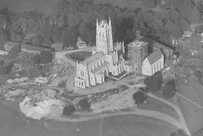This aerial view of the Cathedral shows the Ezekiel Tower fenced with scaffolding. The great stone shed is at the left, the carpenter shop beyond the Council Hall on the right. The architectural studio is hidden by the main tower; the metal shop is off to the left, down the hilly road; and the glass-making shop lies beyond the road glimpsed at upper right. The time is 1924, a date firmly established by the broken pinnacle at the northeast corner of the tower. Struck by lightning following a wedding one evening, the top plunged through the roof and landed where the organist had been playing less than an hour before.
